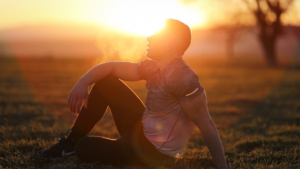 Muscular man sits in rural field at sunset, exhaling visible breath.